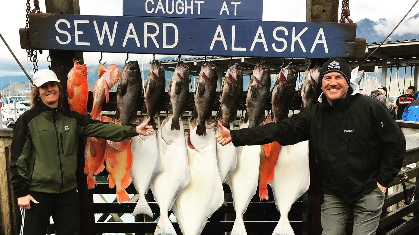 Two anglers beside halibut and rockfish hanging under a sign reading Caught at Seward Alaska